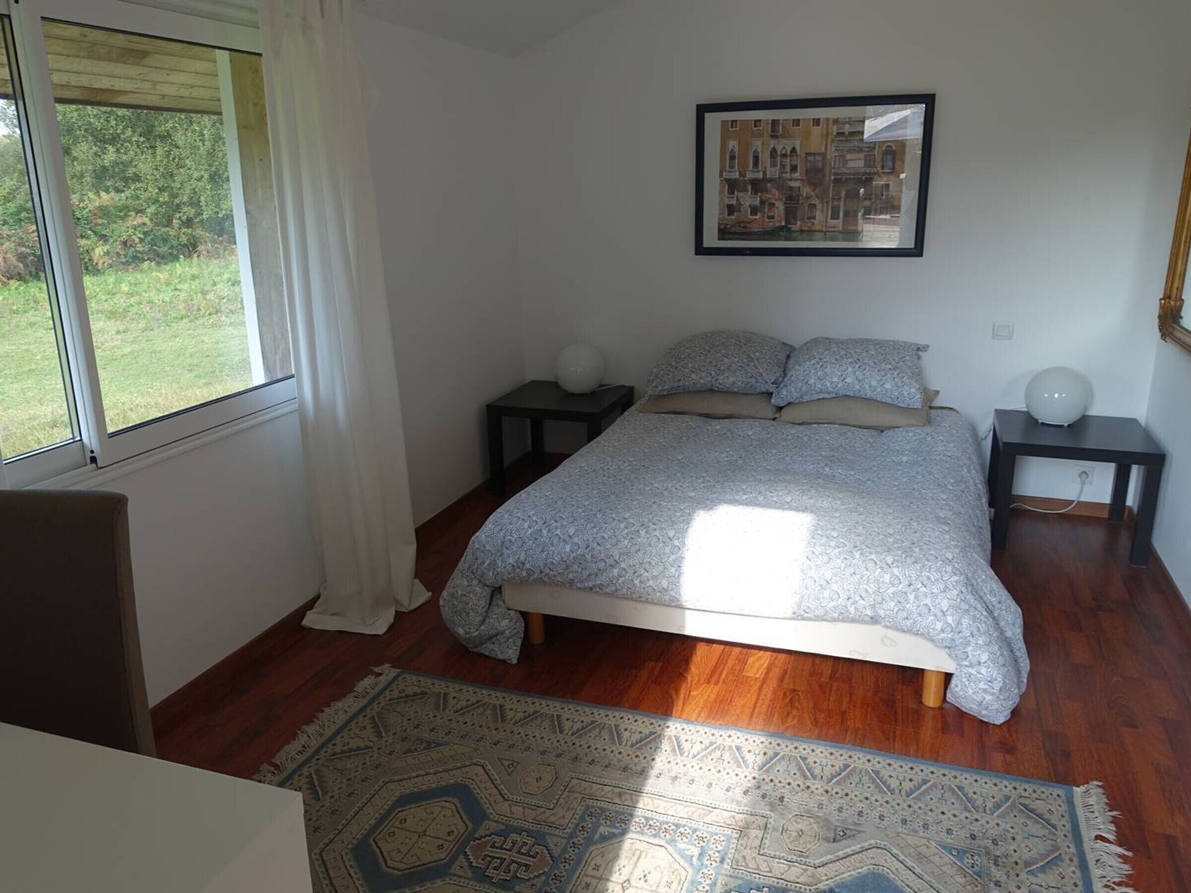Ein Schlafzimmer mit Bett, Nachttisch, Fenster mit Vorhängen und einem gerahmten Bild an der Wand.