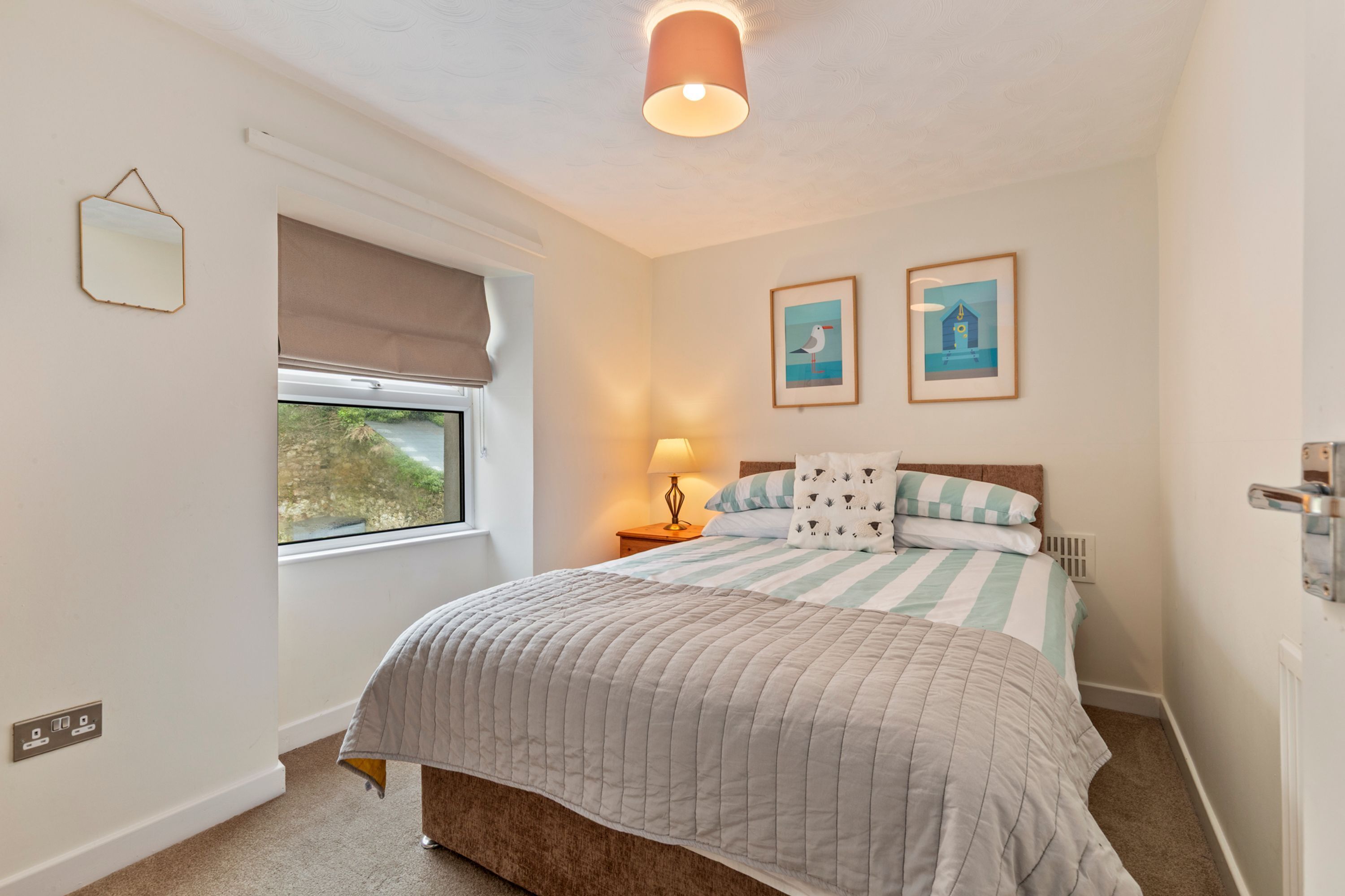 A bedroom with a bed, a window with a view, a bedside lamp, and two framed pictures on the wall.