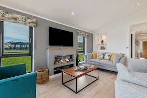 A modern living room with a fireplace, a flat-screen TV, grey sofas, and a view of the outside. - Bryn Hafod - Luxury Lodge Hot Tub Walk to Beach (Tenby)