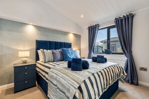 A bedroom with a large bed, a bedside table with a lamp, and a view of the outside through a window with curtains. - Bryn Hafod - Luxury Lodge Hot Tub Walk to Beach (Tenby)
