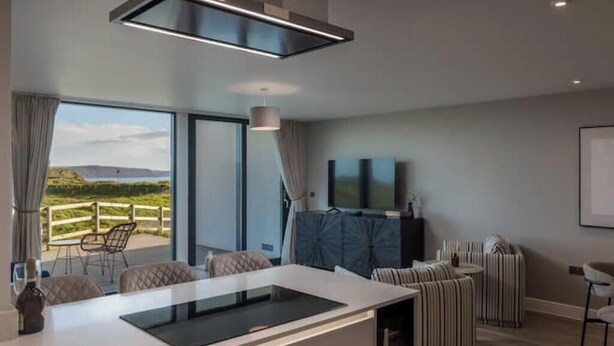 Een moderne keuken met een centraal eiland, een eethoek met gestreepte stoelen, een aan de muur bevestigde televisie en uitzicht op de omgeving.