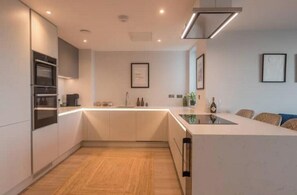 A modern kitchen with built-in appliances, a central island, and a dining area with chairs. - Sea View Stylish Apt with Sauna, Steps to Beach (Bude)