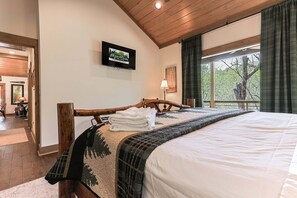 Ein Schlafzimmer mit hölzernem Bettgestell, einem an der Wand befestigten Fernseher, einem Fenster mit Vorhängen und Blick auf Bäume.