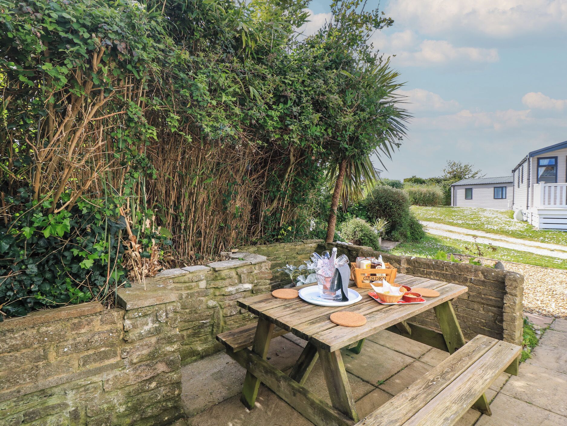 A wooden picnic table set up in a garden with a stone wall, surrounded by lush greenery and a small house in the background.