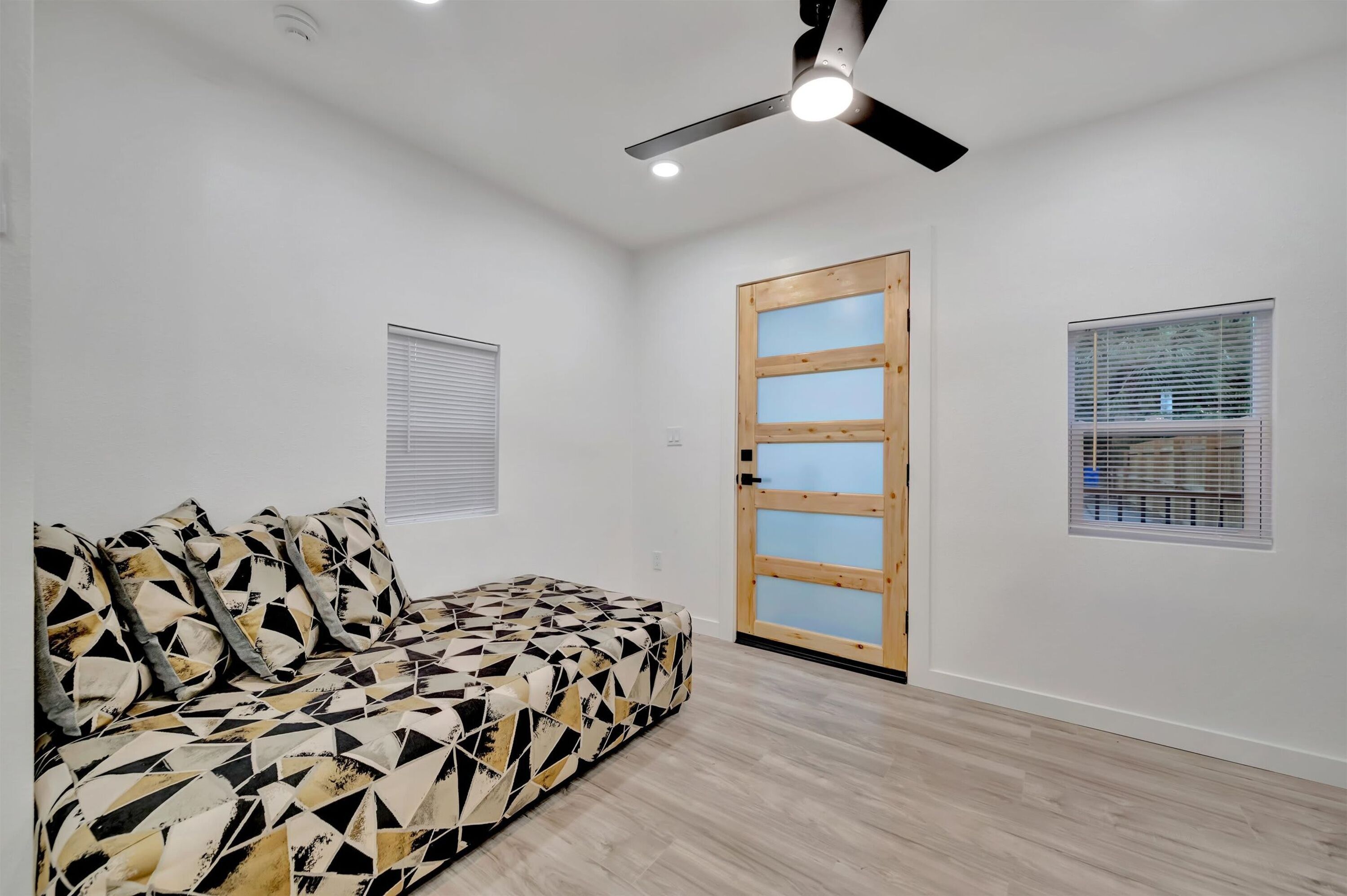 A room with a geometric-patterned sofa, a ceiling fan, a wooden door with glass panels, and two windows with blinds.