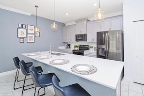 Une cuisine moderne dotée d’un grand îlot central, de tabourets de bar, de luminaires suspendus et d’appareils électroménagers en acier inoxydable.