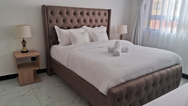 Una cama bien hecha con cabecera capitonada, dos lámparas de noche y una toalla enrollada sobre la cama.