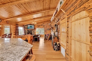 A log cabin interior with a stone fireplace, wooden dining table, and a mounted deer head.