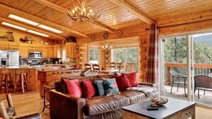 A cabin interior with a wooden dining area, a sofa with cushions, a kitchen with appliances, and a large window overlooking a forest. - Aspen Bear Retreat | Spa, Mountain Views, Near Bear Mountain & Slopes Sleeps 8 (Big Bear City)