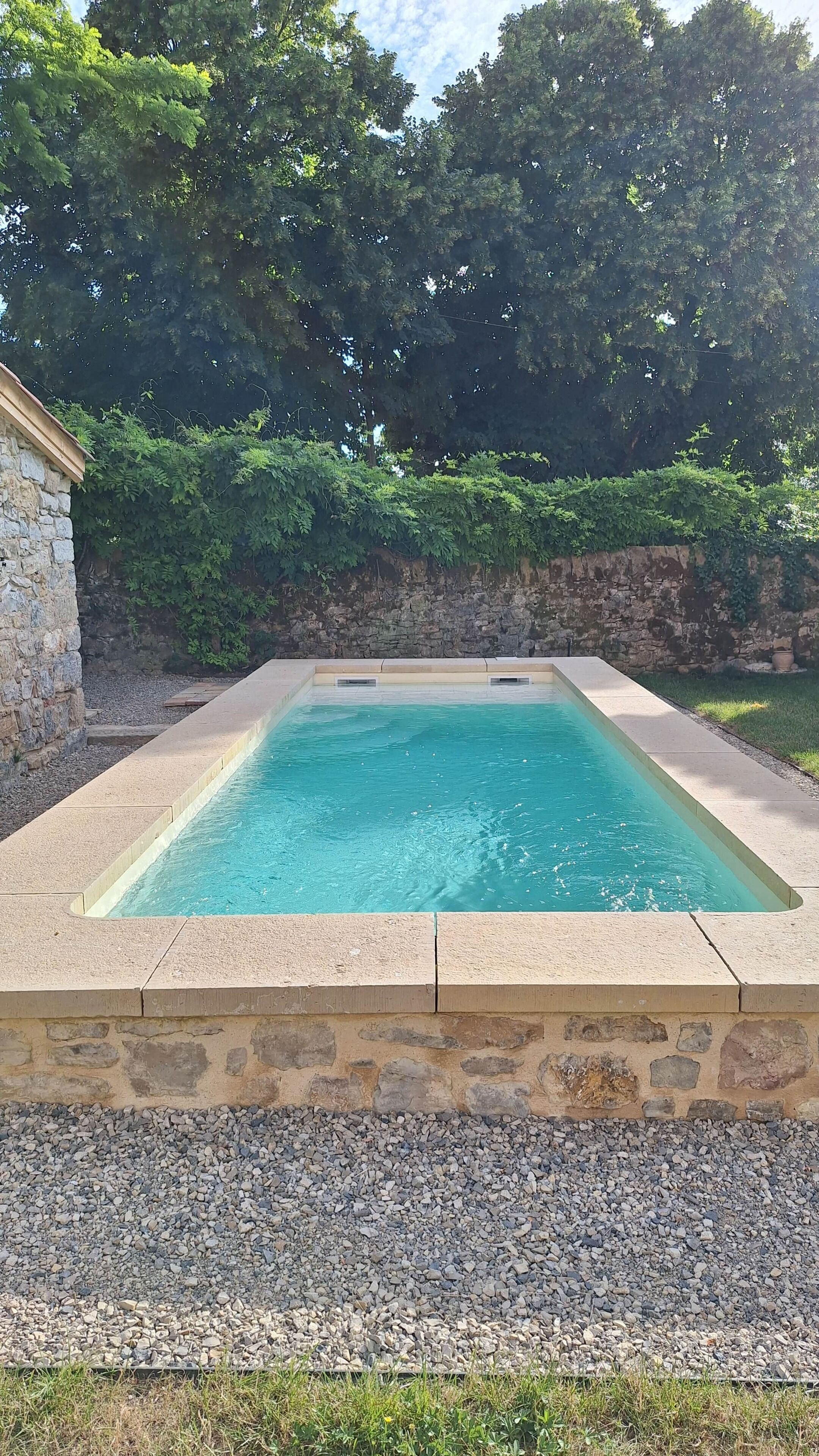 A rectangular swimming pool with a stone border, surrounded by a stone wall and a grassy area.
