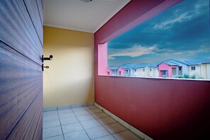 Een kamer met betegelde vloer, een houten deur, een gele wand en een rode wand met uitzicht op huizen.