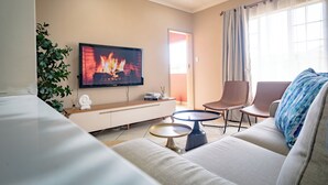 A modern living room with a flat-screen TV, a sofa, two side tables, a potted plant, and a view of the outdoors.