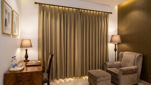 Hotelkamer met een groot raam, een houten bureau met lamp, een stoel, een bank en een klein pouf.