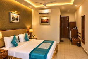 Quarto de hotel com cama, mesas de cabeceira, luminária, ar condicionado, ventoinha de teto, porta e um banco.