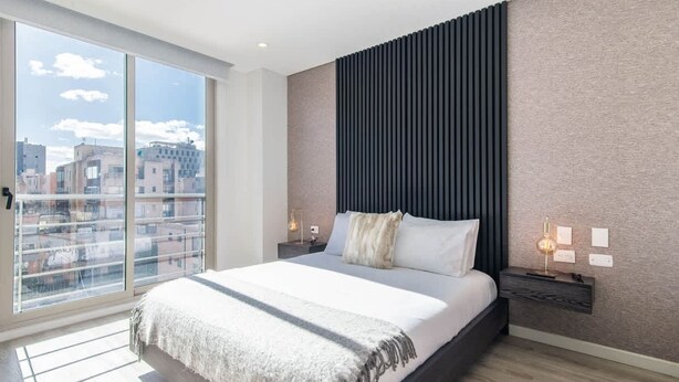 A modern bedroom with a large bed, a city view, and a textured wall. - NOK Cozy 2 BR at Usaquen (Bogotá)