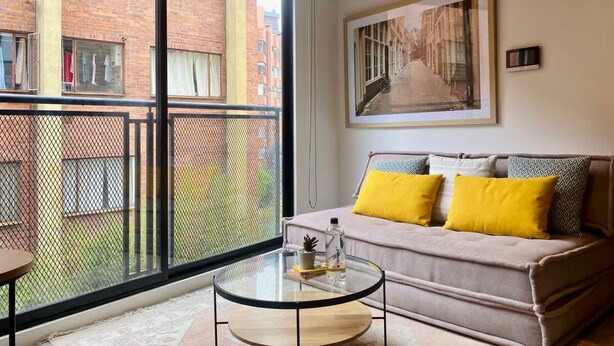 Una sala de estar moderna con un sofá, una mesa de centro de vidrio y un amplio ventanal con vistas a un edificio de ladrillo.