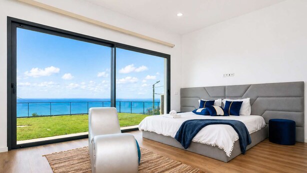 Quarto com uma janela grande, cama, cadeira e uma mesinha redonda.