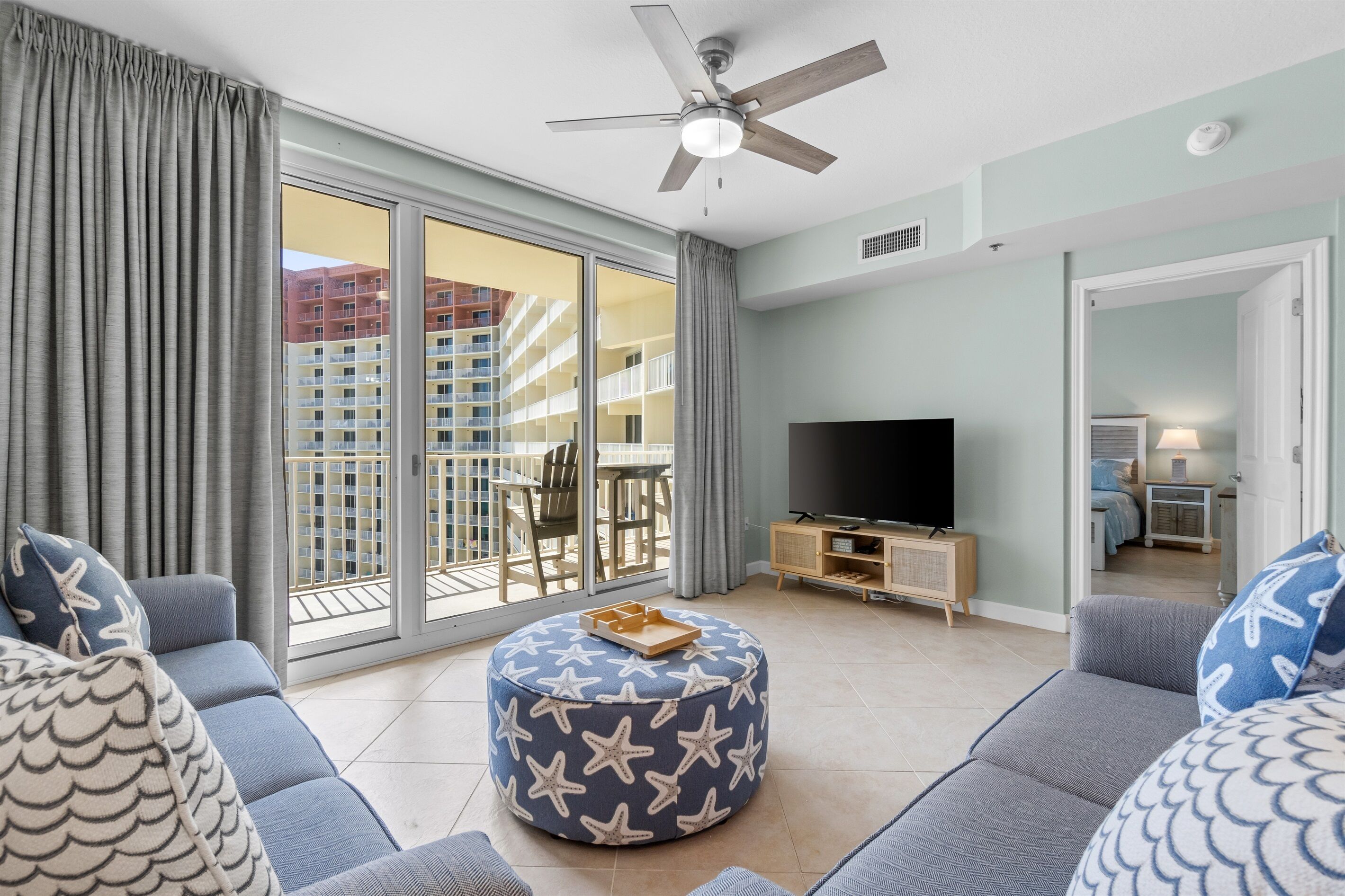 A living room with a sofa, ottoman, TV, and balcony with sliding doors.