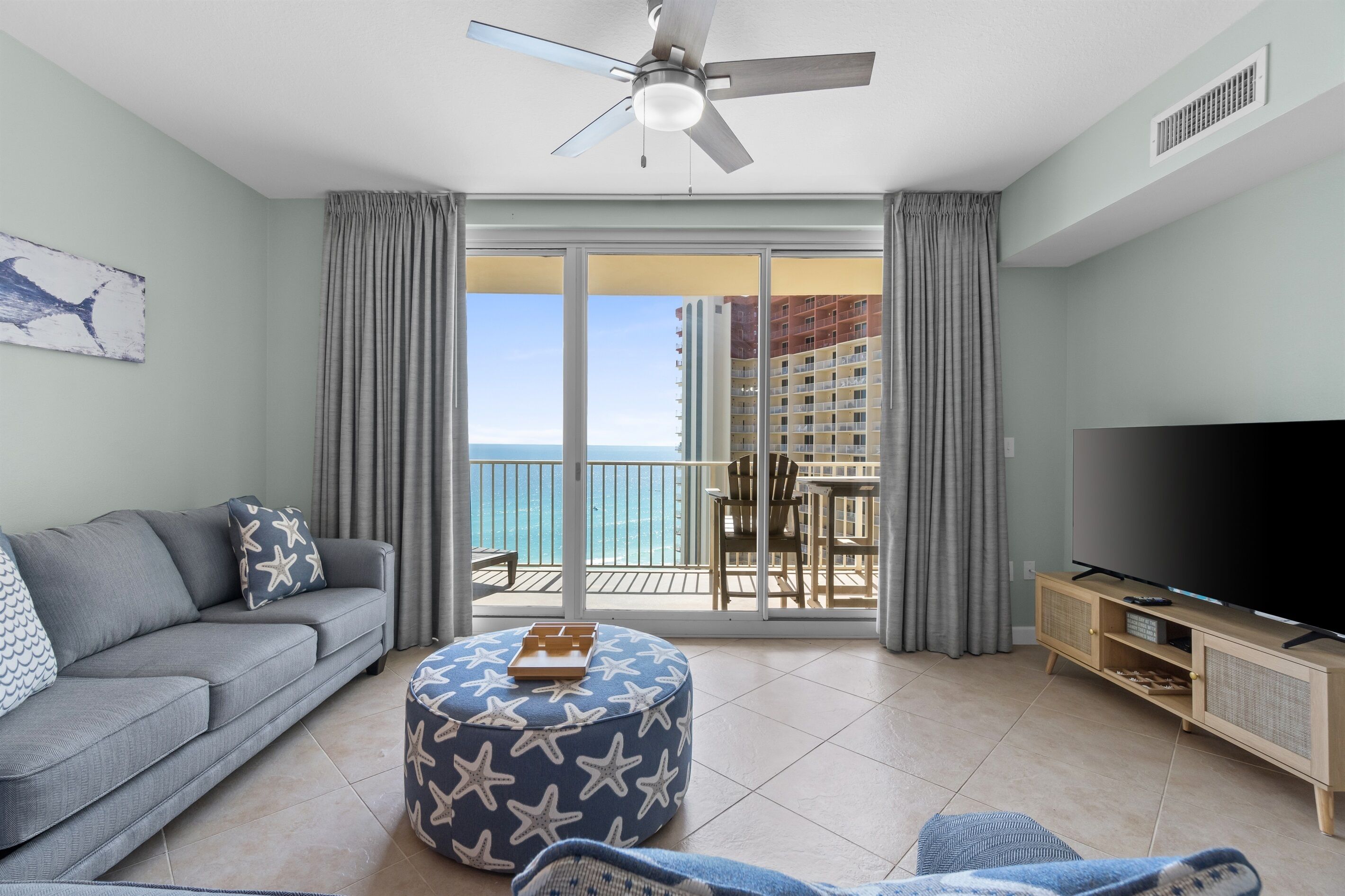 Ein Wohnzimmer mit Sofa, Ottomanen, Fernseher und einem Balkon mit Blick aufs Meer.