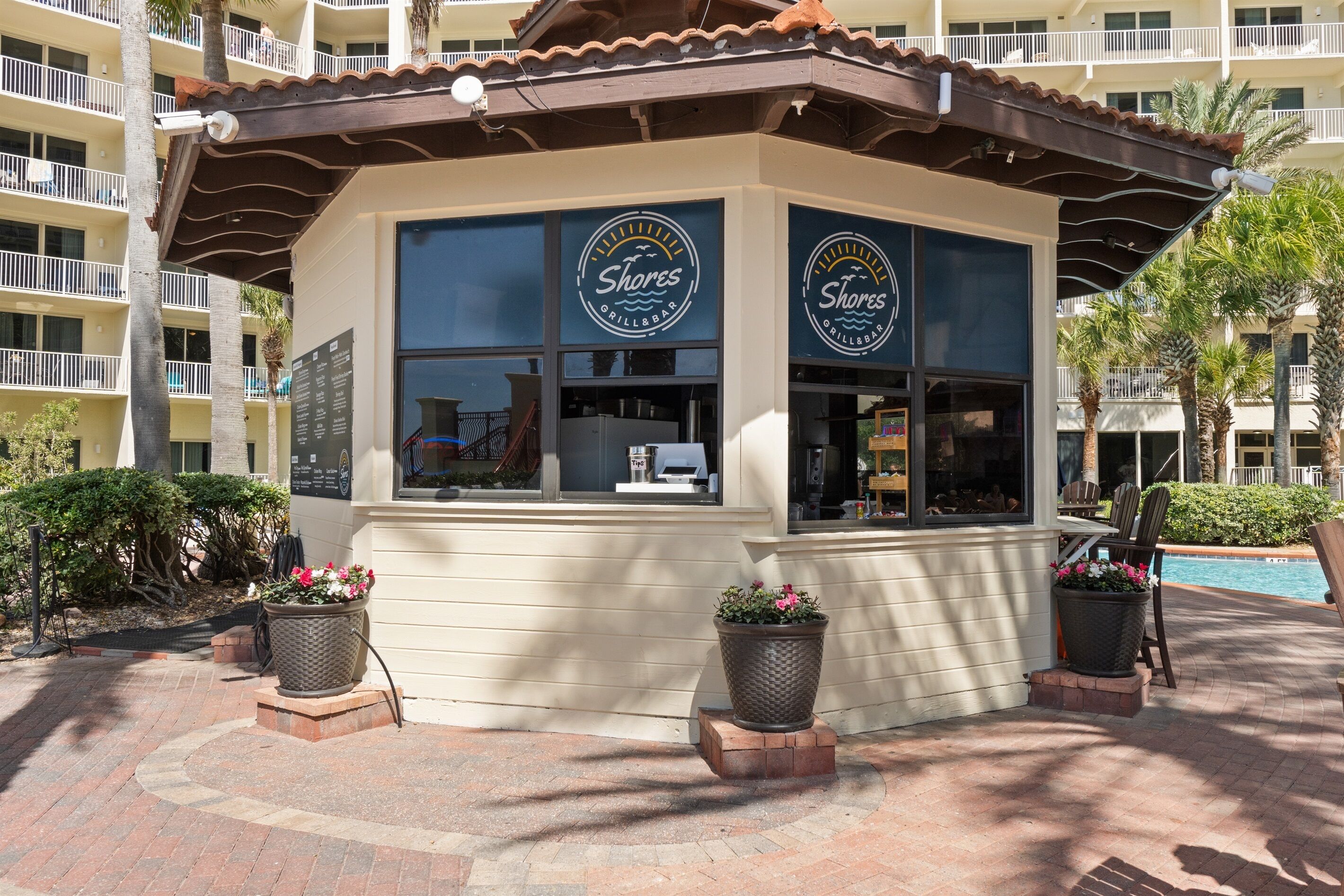 A Shores Grill Bar with a patio area, potted plants, and a pool in the background.