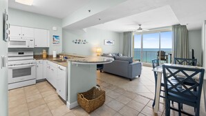 Eine moderne Küche mit weißen Geräten, ein Essbereich mit blauen Stühlen und ein Wohnzimmer mit einem grauen Sofa.
