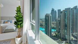 A modern bedroom with a large window, a potted plant, a bed with patterned pillows, and a cityscape view. - Gala Guests - 29 Boulevard Residences (Dubai)