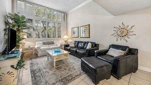 A living room with a sofa, ottoman, coffee table, TV, and a large window with blinds. - Oceanside with Pool, Hear the waves! Beach Walker Villas (Fernandina Beach)