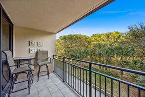 A balcony with two chairs and a table, a railing, and a view of trees and shrubs. - Oceanside with Pool, Hear the waves! Beach Walker Villas (Fernandina Beach)