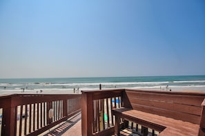 Eine Holzterrasse mit Blick auf einen Strand, wo sich Menschen im Wasser und im Sand aufhalten.
