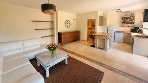 A living room with a sofa, a coffee table with flowers, a dining area with a kitchen in the background. - Charming Private Cottage with Garden & Parking in Bar-sur-Loup (Le Bar-sur-Loup)