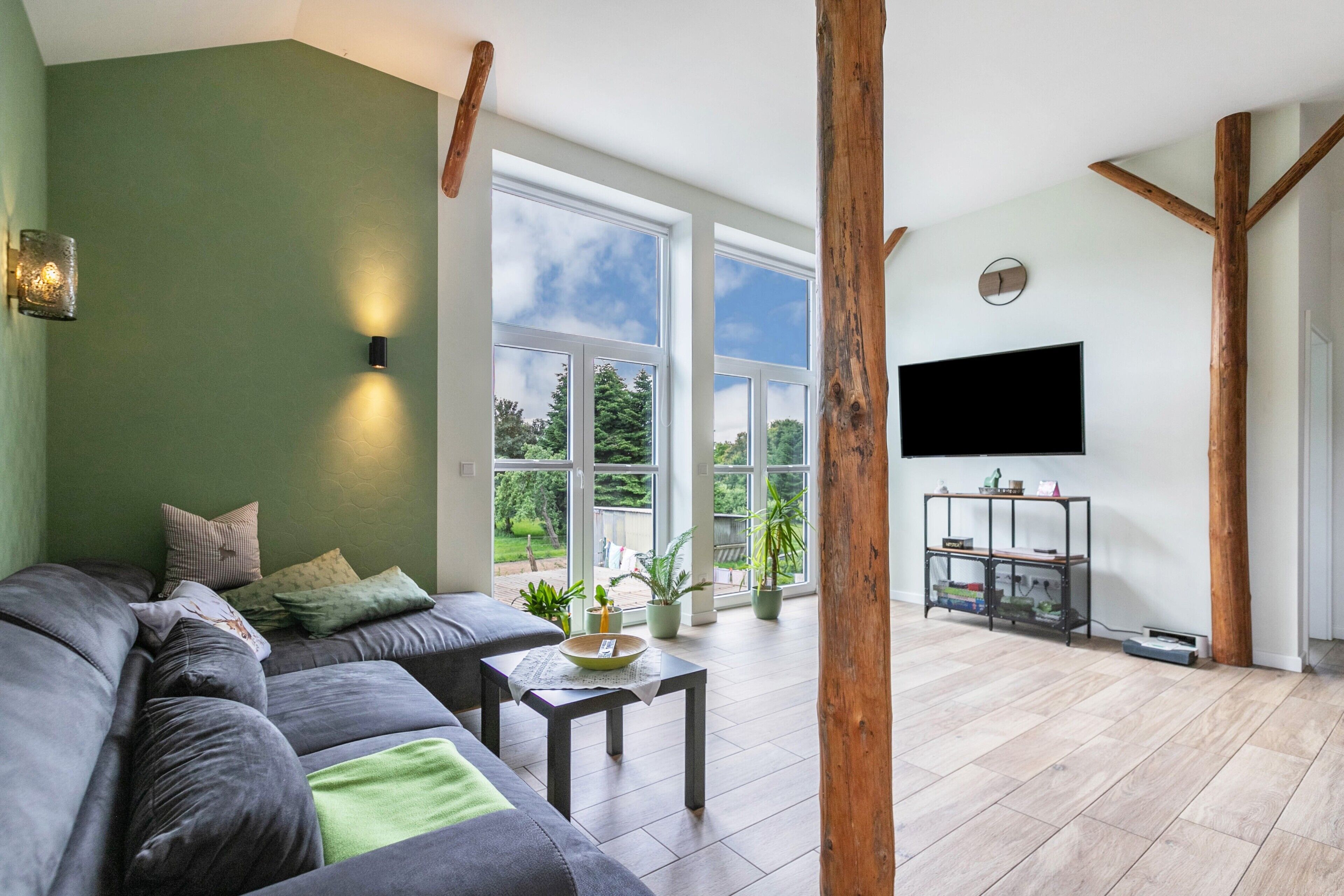 Ein modernes Wohnzimmer mit grauem Sofa, einem Couchtisch und einem an der Wand befestigten Fernseher. Große Fenster sorgen für natürliches Licht und Ausblicke ins Freie.