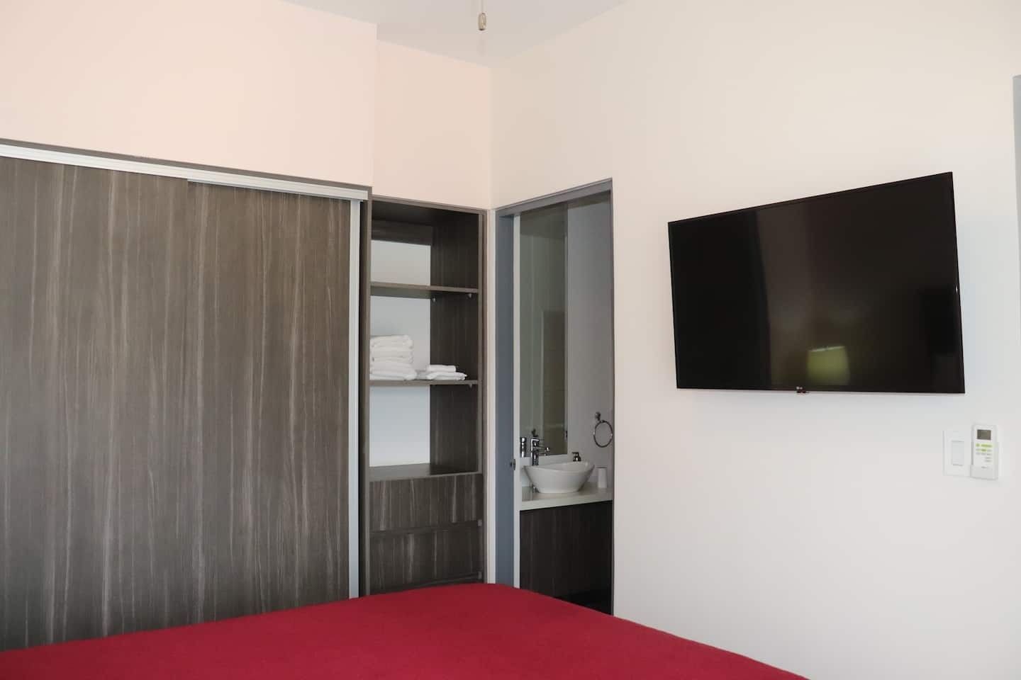 A bedroom with a red bed, a wardrobe, a built-in shelf with folded towels, a wall-mounted TV, and a small bedside table with a lamp.