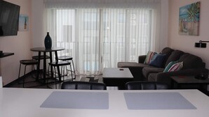 A living room with a sofa, a dining table, a small kitchen area, and a large window with sheer curtains. - Great apartment near the Airport with Air Condit. (Alajuela)