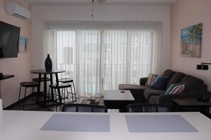 Una sala de estar con sofá, una mesa de comedor, una pequeña cocina y un ventanal con cortinas translúcidas.