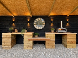 A wooden outdoor seating area with a clock, a pizza oven, and potted plants.