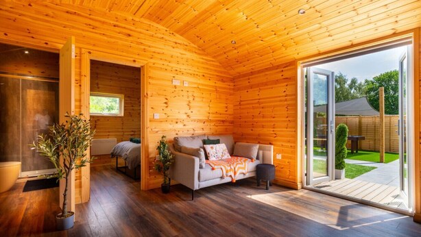A log cabin interior with a sofa, a potted plant, and a sliding glass door leading to a garden. - The Cabin  @The Outside Inn (Mountfield)