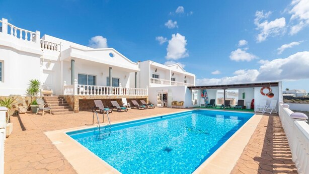 Une piscine avec des chaises longues, un bâtiment blanc doté de balcons et un ciel d’un bleu limpide.