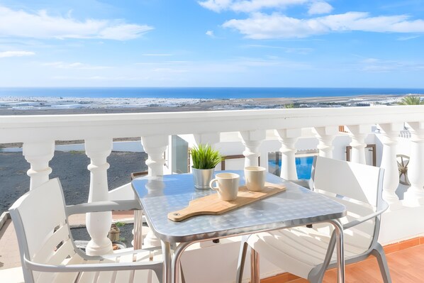 A balcony with a table, chairs, a plant, and a view of the beach and ocean.