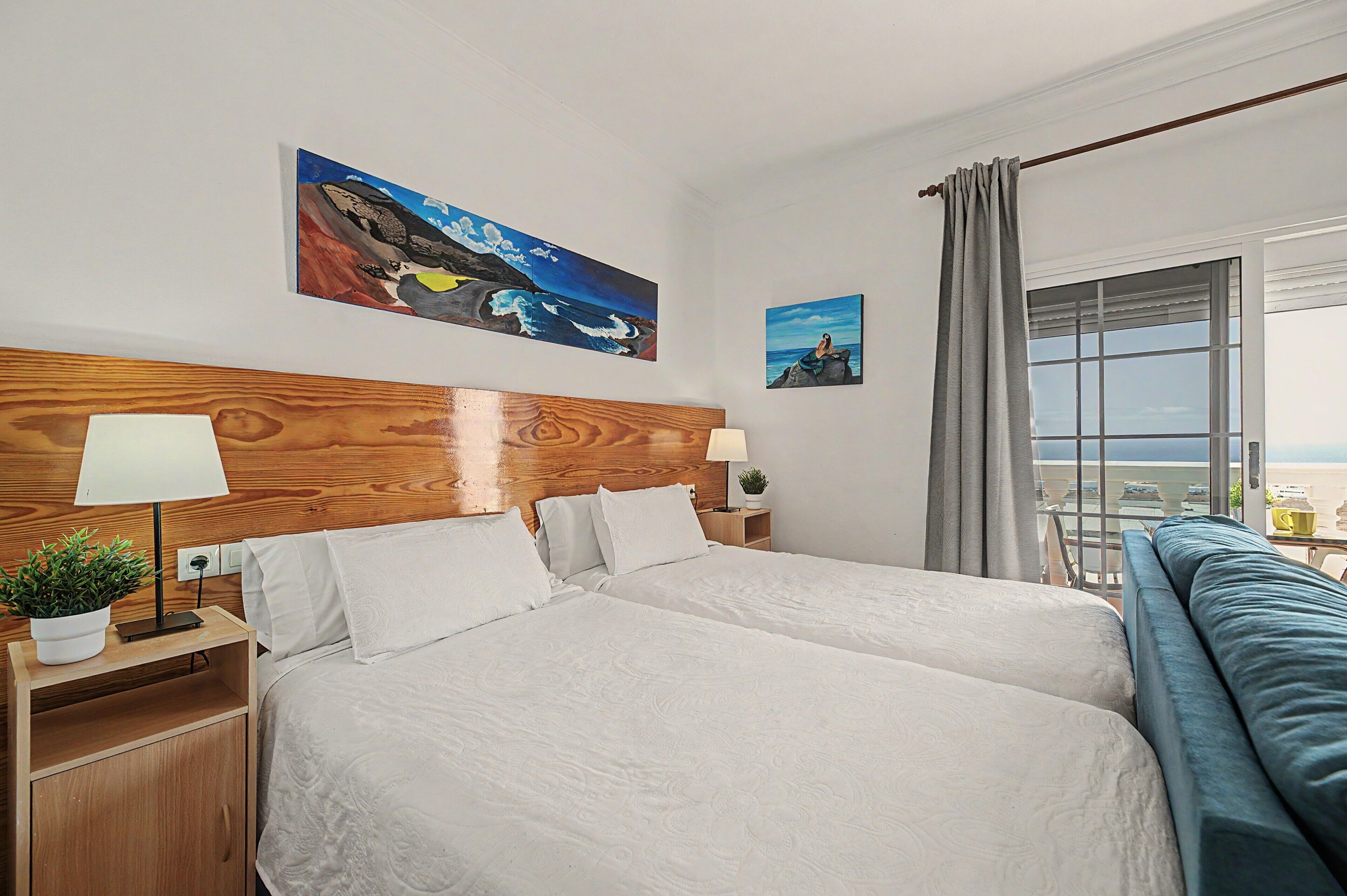 Une chambre d’hôtel avec un grand lit, deux tables de chevet, une petite table d’appoint, une plante en pot et une vue sur la mer par la fenêtre.