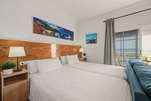 Een hotelkamer met een groot bed, twee nachtkastjes, een plant in pot en uitzicht op de zee door een raam.