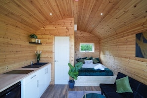 Une petite cabane en bois comprenant un coin cuisine, un lit avec une literie verte et une petite fenêtre.