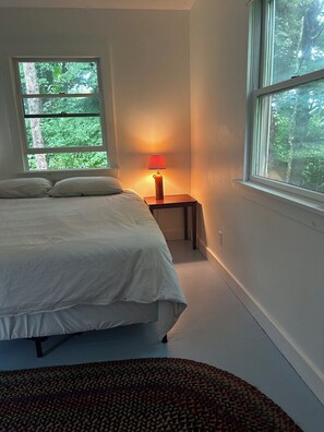 Ein Schlafzimmer mit Bett, Nachttisch mit Lampe, Fenster mit Blick auf Bäume und einem gemusterten Teppich.