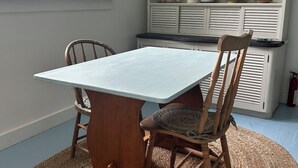 A dining area with a table and chairs, a kitchen with shelves, and a window with a view of a house. - Cape Rosier Cozy Guesthouse (Harborside)