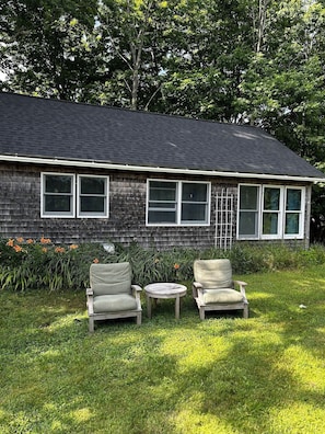 A house with a dark shingled roof, a front porch with two chairs and a small table, and a garden with flowers and grass.