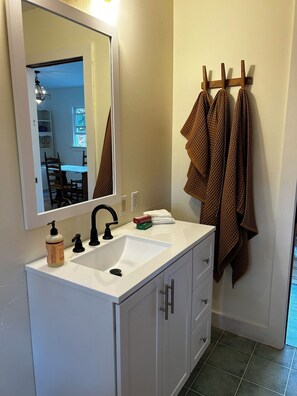 A bathroom with a white sink, a mirror, a towel rack with towels, and a view into a dining area.
