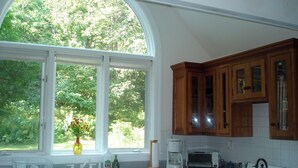 A kitchen with a large window, wooden cabinets, a stove, and a coffee maker. - Hearthstone Cottage , Private cottage on Waters Edge! (Blue Hill)