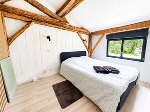 A bedroom with a bed, a window, wooden beams, and a view of greenery. - Les cabanes (Noaillan)