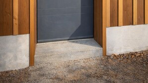 A modern building entrance with a dark door, wooden paneling, and a concrete step. - Les cabanes (Noaillan)