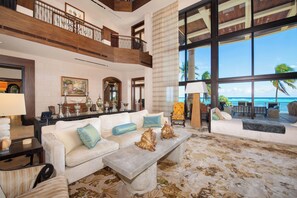 A spacious living area with a high ceiling, a balcony overlooking the ocean, and a large window providing a view of the sea. - Castillo Caribe: Ultra-Luxe Estate w/Pool Grotto, Tennis & Pickle Ball Courts + Home Spa & Gym (South Sound)