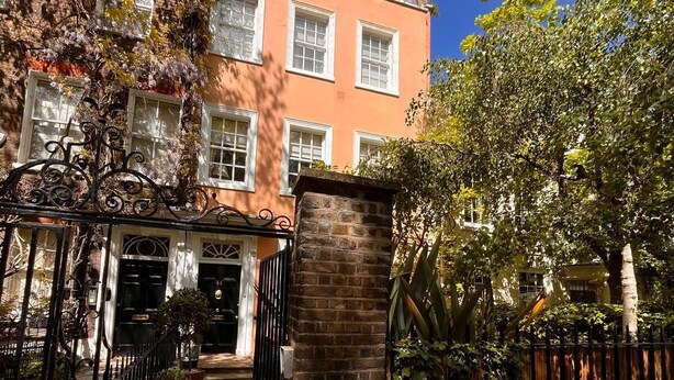 Uma entrada com portão e um pilar de tijolos, um caminho que leva a um edifício com várias janelas, e uma cerca com árvores.